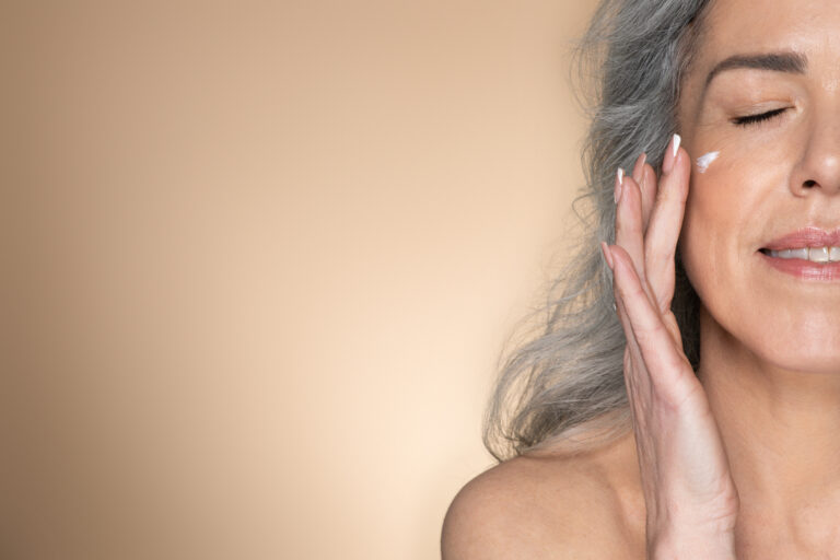 Half face of senior woman applying anti-aging or moisturising face cream, posing on beige studio background, free space